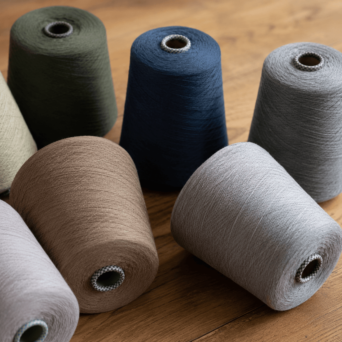 Industrial cone yarns arranged casually on a warm wood wax oil table, photographed from above in soft natural studio light. White, cream, beige, and deep navy yarn cones create a quiet, textural composition with subtle shadows and realistic fiber detail.