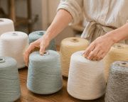 Hands arranging muted cone yarns on a wooden studio table, featuring soft mint, cream, pale yellow, heather gray, and brown industrial yarn cones with realistic fiber texture, photographed in a warm shallow-depth workshop setting. Conceptual image · AI-as