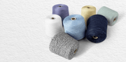 Soft-toned industrial cone yarns arranged casually on a textured white paper backdrop, featuring lavender, cream, pale mint, sky blue, navy melange, and marled gray yarn cones with visible fiber texture and realistic winding details. Quiet editorial produ