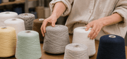 Hands arranging muted cone yarns on a wooden studio table, featuring soft mint, cream, pale yellow, heather gray, and brown industrial yarn cones with realistic fiber texture, photographed in a warm shallow-depth workshop setting. Conceptual image · AI-as